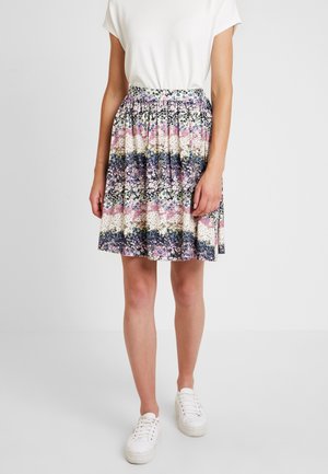 Personne portant un haut blanc à manches courtes, une jupe fleurie multicolore longueur genoux et des baskets blanches, debout devant un fond uni.