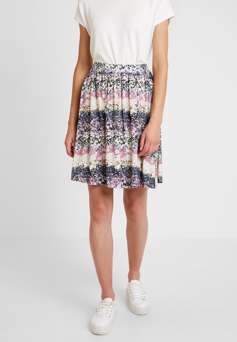 Personne portant un haut blanc à manches courtes, une jupe fleurie multicolore longueur genoux et des baskets blanches, debout devant un fond uni.
