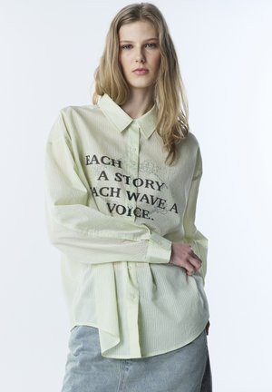 Jonge vrouw met lang blond haar, gekleed in een lichtgele gestreepte blouse met zwarte tekst en een lichte denim rok, tegen een witte achtergrond.