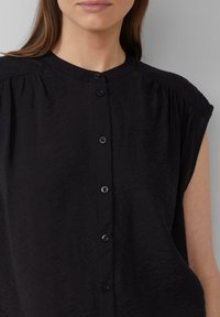 Blouse noire à manches courtes avec un tissu texturé, dotée d'un col haut et d'une fermeture à boutons à l'avant avec cinq boutons noirs.