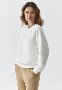 Biely sweatshirt s okrúhlym výstrihom, dizajnom so spustenými ramenami a rebrovanými manžetami. Spárované s béžovými nohavicami vo voľnom strihu.