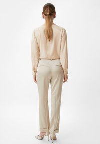 Kvinde med langt hår sat i en lav hestehale iført en beige bluse, matchende lige ben bukser og højhælede sko, stående med ryggen til.