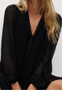 Blusa negra plisada con mangas largas transparentes, escote en V y corte holgado. La tela tiene una textura ligera y fluida.