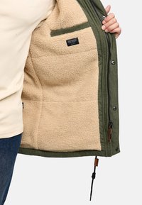 Grüne Außenjacke mit einem weichen beigen Fleece-Futter, ausgestattet mit einem Reißverschluss, Seitentaschen und einem Etikett innen. Verstellbarer Saum mit Kordelzug.