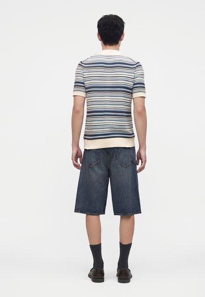 Homme debout, de dos, portant un pull rayé à manches courtes, un short en jean foncé au genou, des chaussettes noires et des chaussures noires sur un fond uni.