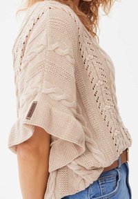 Suéter de punto beige con mangas anchas, que presenta un patrón de trenzas y puntadas abiertas. Etiqueta presente en la manga. Llevado con pantalones cortos de mezclilla azules.