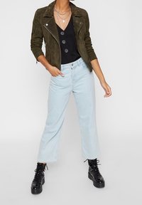 Oliwkowa zamszowa kurtka motocyklowa z srebrnym zamkiem, czarna koszula z guzikami, jasnoniebieskie spodnie z szerokimi nogawkami i czarne masywne buty na sznurówki.