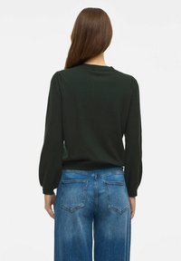 Top cu mâneci lungi, de culoare verde închis, cu guler rotund și umeri bufanți, asortat cu blugi lejeri, de talie înaltă, de culoare albastru deschis.
