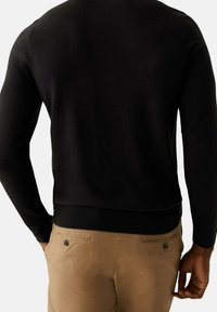 Pull en maille noire avec une coupe ajustée et un ourlet côtelé, porté avec un pantalon chino beige. Comprend des manches longues et une finition à texture lisse.