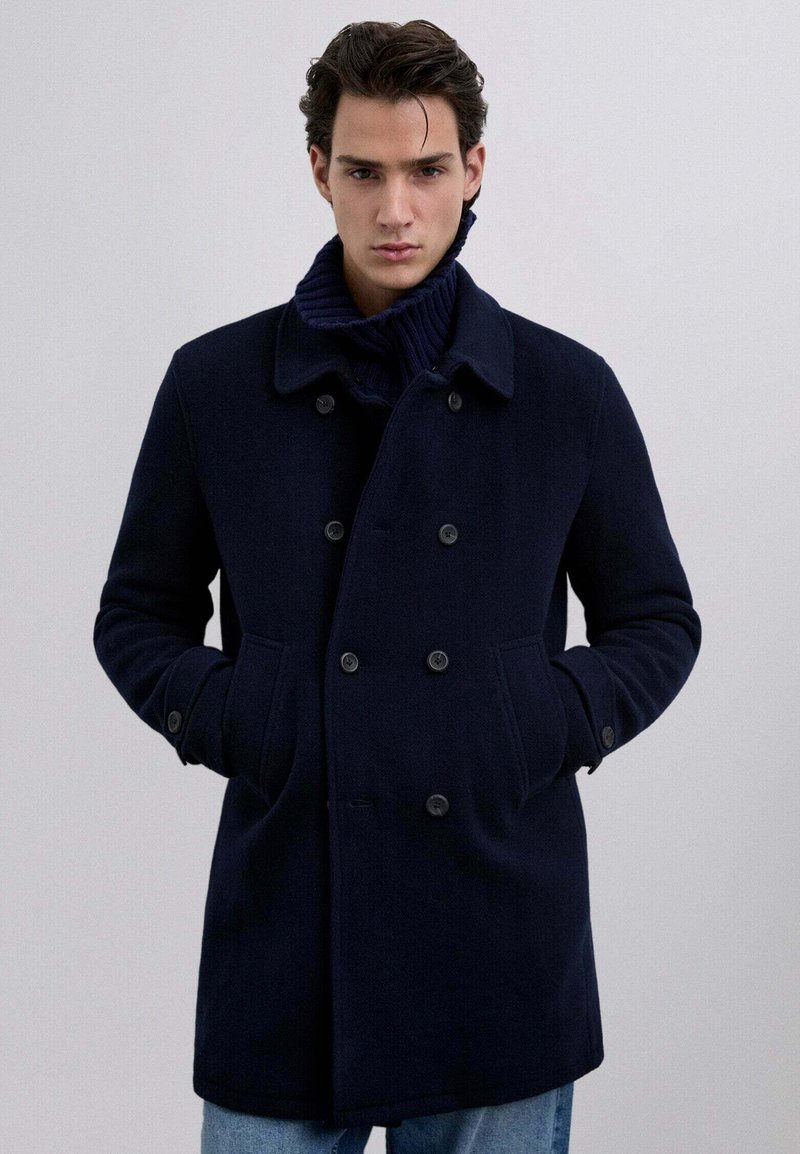 Marineblå uldblandingsfrakke med klapkrave, dobbeltknaplukning, sidelommer og ribstrikket kravedetalje.