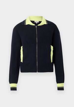 Naisel seisab, kandes musta lukuga kampsunit kollaste detailidega, tumedasiniseid teksapükse ja pruune lainesid jalatseid heledal taustal.