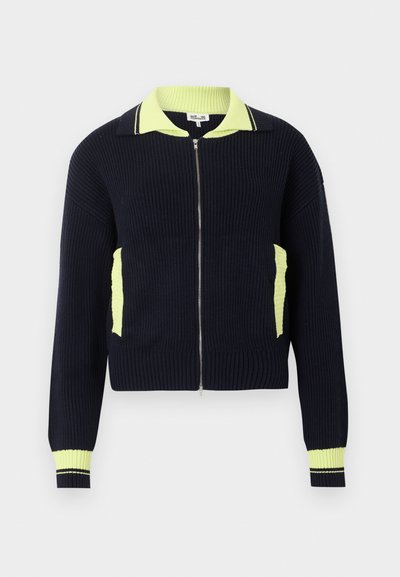 Πλεκτό ζακέτα με φερμουάρ σε ναυτικό μπλε χρώμα, με ρίγες, γιακά σε νέον κίτρινο, άκρες τσεπών και τελειώματα μανικιών, εμφανίζεται πάνω σε απλό ανοιχτό φόντο.