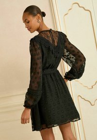 Rochie neagră cu model cu buline, mâneci lungi din material transparent, detalii din dantelă la decolteu și spate, talie strânsă și tiv evazat.