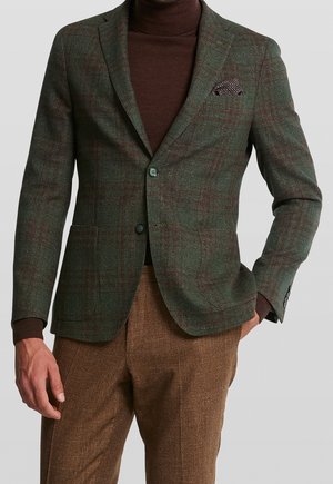 Mann trägt grünen karierten Blazer über braunem Rollkragenpullover und braunen strukturierten Hosen, mit sichtbarem kariertem Einstecktuch.