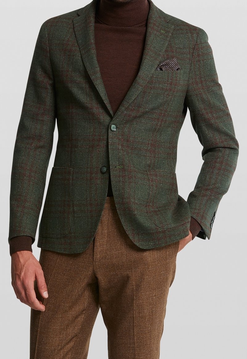Homme portant un blazer à carreaux verts sur un col roulé marron et un pantalon marron texturé, avec un mouchoir à carreaux visible.