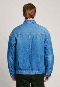 Chaqueta de mezclilla azul con corte clásico, que presenta un cuello con punta, un yugo en la parte posterior y detalles de costura a lo largo de las costuras. Mangas largas con puños abotonados.