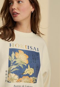 Valkoinen collegepaita, jossa on Hokusai-printti pionista ja kanarialinnusta, sinisellä ja oranssilla korostuksella sekä teksturoitua kangasta.