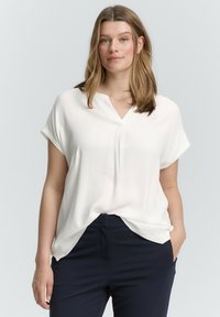 Blouse blanche à manches courtes avec un col en V et une coupe décontractée, fabriquée en tissu lisse. Associée à un pantalon ajusté bleu marine.