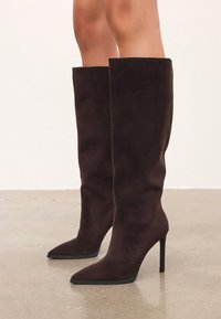 Knee-high tmavohnedé semišové čižmy s špicatými prstami a stiletto podpätkami nosené na holých nohách na neutrálnom pozadí podlahy.