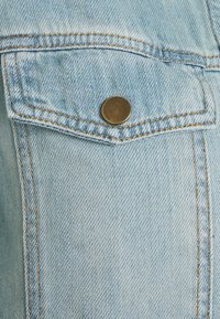Helle blaue Denim-Stoff mit einer Klappen-Tasche, die durch einen runden Metallknopf gesichert ist, und sichtbaren orangefarbenen Nähten.