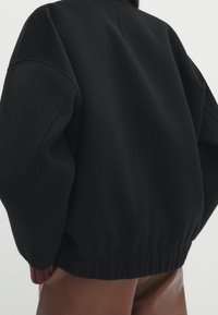 Veste en polaire noire à coupe décontractée, avec des poignets et un ourlet côtelés, présentant une subtile touche de rouge en bas. Matière texturée et douce.