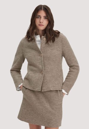 Frau trägt ein beigefarbenes Wollblazer mit drei Knöpfen und passendem knielangem Rock, Hände in den Taschen, vor einfachem Hintergrund.