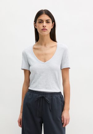 Femme portant un t-shirt à manches courtes gris clair avec un col en V et un pantalon gris foncé à cordon de serrage, debout devant un fond blanc uni.
