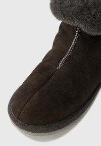 Καφέ σουέτ μπότες με γκρι γούνινη μανσέτα, με κεντρική ραφή και ραφές αντίθετου χρώματος κατά μήκος της άκρης. Διακρίνεται η λεπτομέρεια της υφασματοποιημένης σόλας.