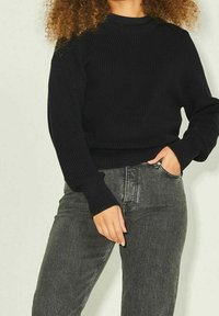 Maglione nero lavorato a maglia con texture a costine, scollo tondo e maniche lunghe, abbinato a jeans grigi scuri a vita alta.