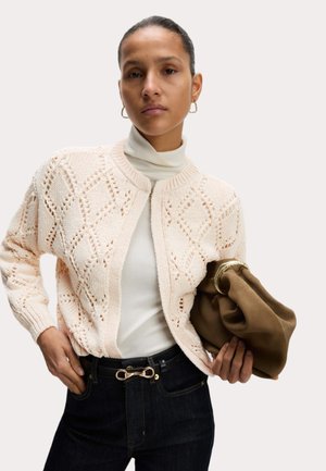 Lichtroze gebreide cardigan met diamantpatronen, gedragen over een witte coltrui. Donkere jeans met een unieke gouden gesp. Bruine clutch tas in de hand.