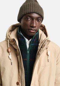 Beżowa kurtka z kapturem, z akcentami ze skóry i zapięciem na zamek. Warstwowa zielona kurtka polarowa z wzorem w kratę pod spodem. Oliwkowy dziany beret.