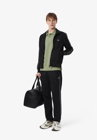 Giacca nera con zip e strisce bianche, polo verde, pantaloni neri e borsa a tracolla nera. Le sneakers presentano una combinazione di bianco e verde.