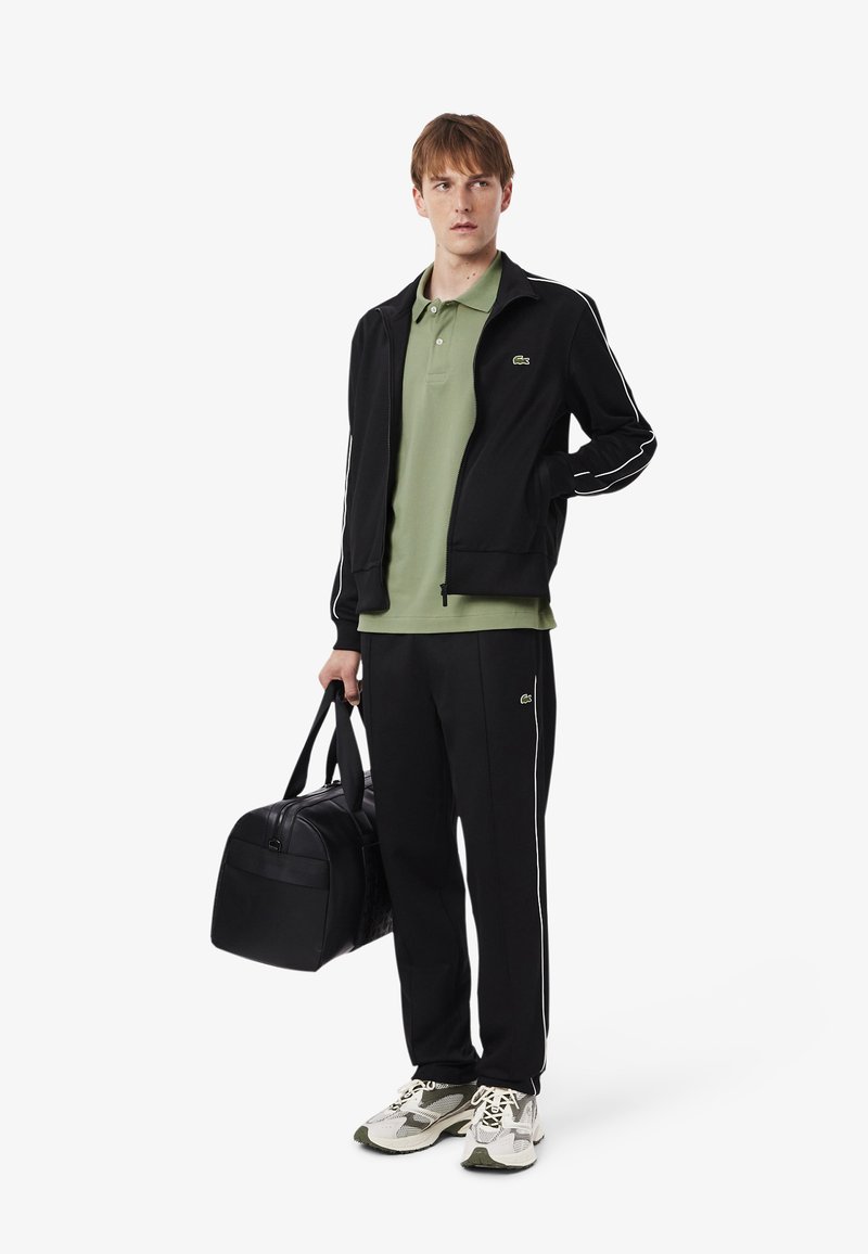 Black zip-up jacket with white stripes, green polo shirt, black pants, and a black duffel bag. Sneakers feature a mix of white and green.