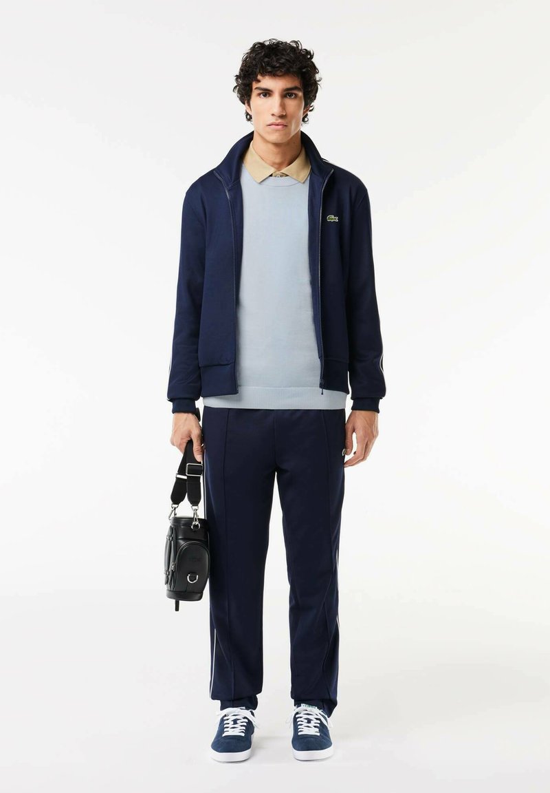 Giacca e pantaloni della tuta navy con polo blu chiaro, rifiniture bianche e sneaker abbinati. Il modello tiene una piccola borsa nera.