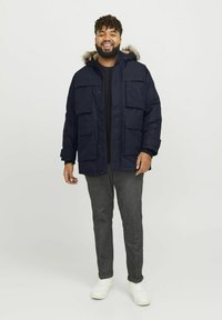 Marine parka met een met bont gevoerde capuchon, ritssluiting en knopen, meerdere voorkruiszakken, gedragen over een zwarte trui en grijze gestructureerde broek.