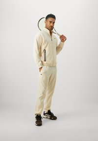 Light cream tracksuit with a zippered jacket and elastic waist pants, featuring black accents, worn with brown sports shoes, holding a tennis racket.