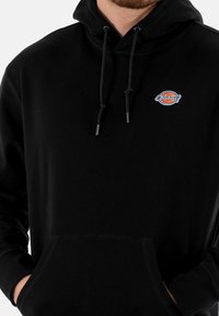 Schwarzer Hoodie aus weichem Material mit einem gestickten Logo auf der Brust, verstellbaren Kordeln und einer Kängurutasche an der Vorderseite.