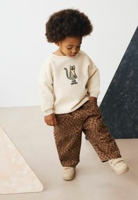 Felpa beige con design a stampa leopardata; pantaloni larghi a fantasia marrone; sneakers crema; postura giocosa del bambino.