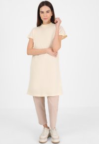 Abito beige al ginocchio con maniche corte e scollo alto, abbinato a pantaloni rosa chiaro a sigaretta e sneaker bianche chunky.