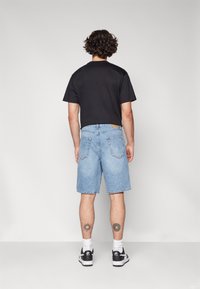 Sort kortærmet T-shirt, lyseblå denimshorts og sorte sneakers. Synlige tatoveringer på benene. Ensfarvet baggrund, fuld kropsvisning fra bagfra.