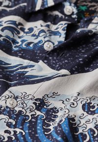 Camicia blu scuro e bianca con motivi a onde oceaniche e bottoni bianchi, con dettagli di schiuma e spruzzi d'acqua.