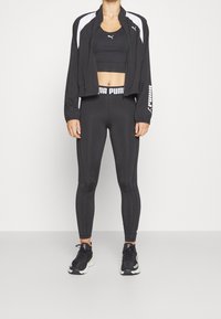 Musta urheilu takki valkoisilla yksityiskohdilla, musta lyhyt toppi ja tiukat legginsit, joissa on logon vyötärö; yhdistettynä mustiin lenkkareihin.