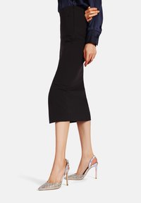 Gonna nera aderente abbinata a sandali argentati con cinturini decorati con strass, caratterizzati da una punta appuntita e dettagli metallici sul tacco.