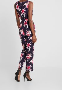 Combinaison fleurie avec un fond bleu marine, des fleurs roses et blanches, sans manches et avec des jambes fuselées. Portée avec des chaussures à talons noirs.