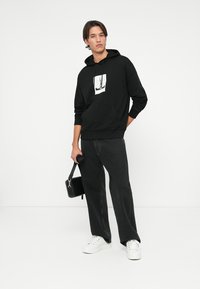 Svart huvtröja med ett grafiskt tryck fram, kombinerad med mörka jeans och vita sneakers. Håller en liten svart väska.
