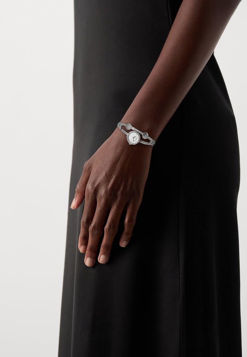 Bras à la peau foncée portant une montre-bracelet argentée avec un cadran rond, reposant contre une robe noire sur un fond clair.