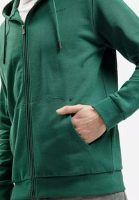 Zielona bluza z zamkiem błyskawicznym, wykonana z teksturowanej tkaniny, z przednimi kieszeniami i regulowanym sznurkiem. Posiada dopasowane mankiety oraz luźny krój.