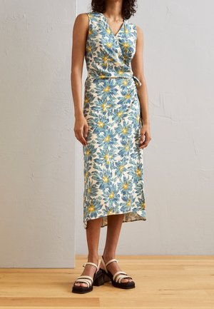 Ærmeløs omslagskjole med blå og gul blomsterprint, midi-længde, båret med beige og sorte blokhælede sandaler på trægulv.