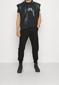 Veste sans manches en cuir noir, t-shirt graphique noir, pantalon cargo noir avec poches, et bottes noires à semelles texturées.