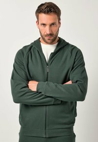 Grüne Zip-Hoodie aus strukturierter Stoff, mit Raglanärmeln und einem Frontreißverschluss. Das Modell hat die Arme verschränkt und trägt ein weißes Shirt darunter.